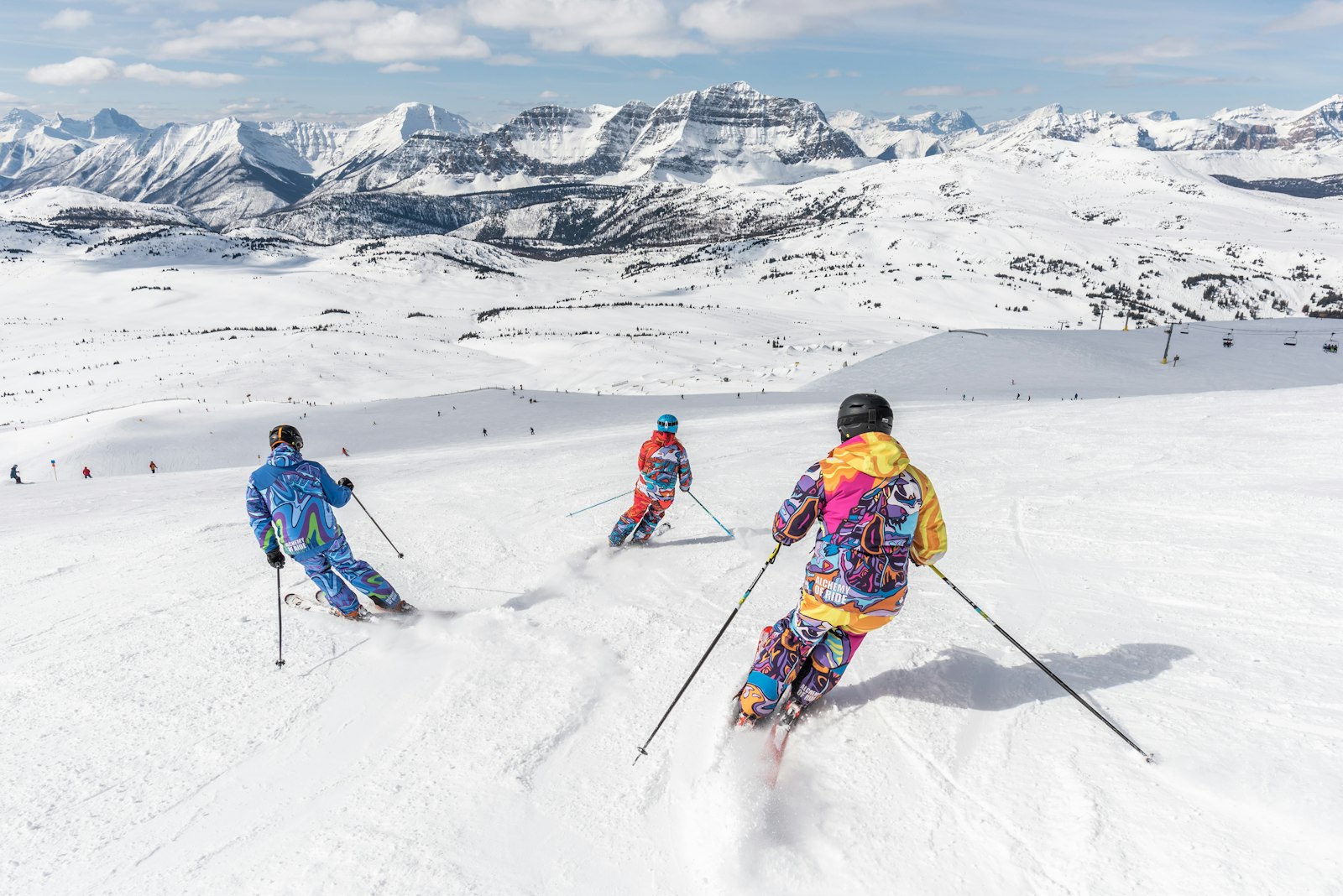 Three skiers on a wide, sunlit alpine slope, with a high mountain range beyond.